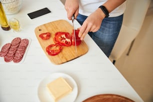 Close up of a woman cutting a red bell pepper on a wooden board. Sausages and butter in front of her
