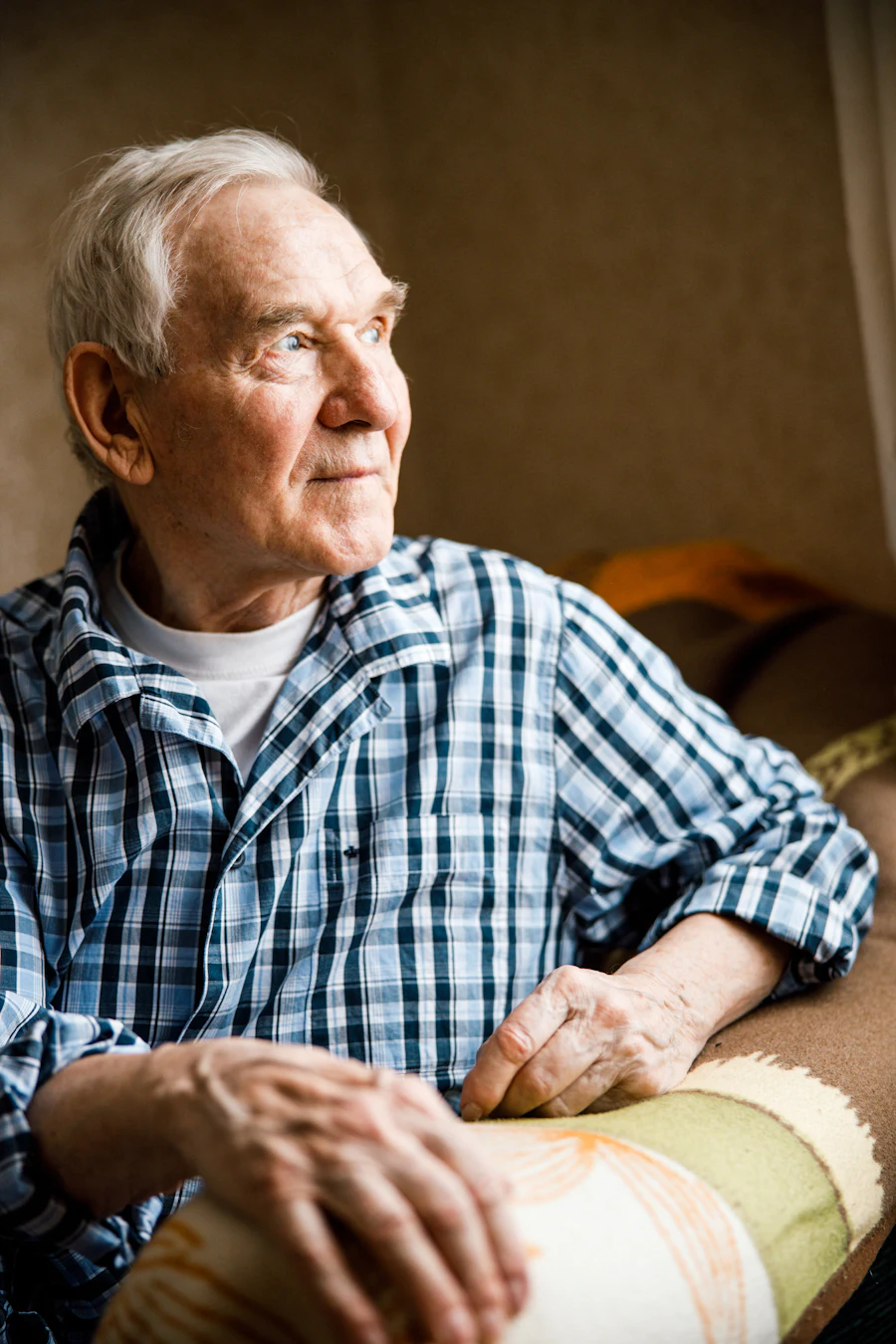 An older man in a checked shirt sitting by a window in a softly lit room