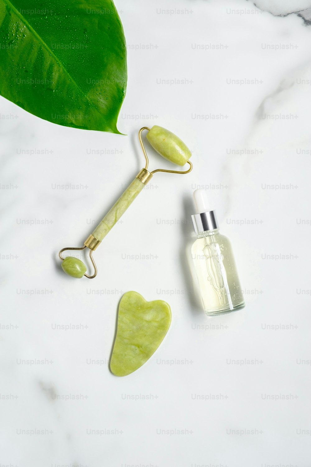 Jade roller with natural anti aging jade stone, essential oil dropper bottle and green leaf on marble table. Facial skin care concept. Flat lay, top view.