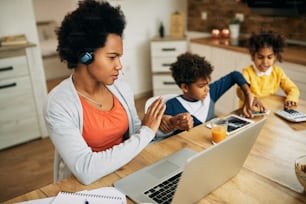 African American working mother silences her children while having conference call over laptop at home.