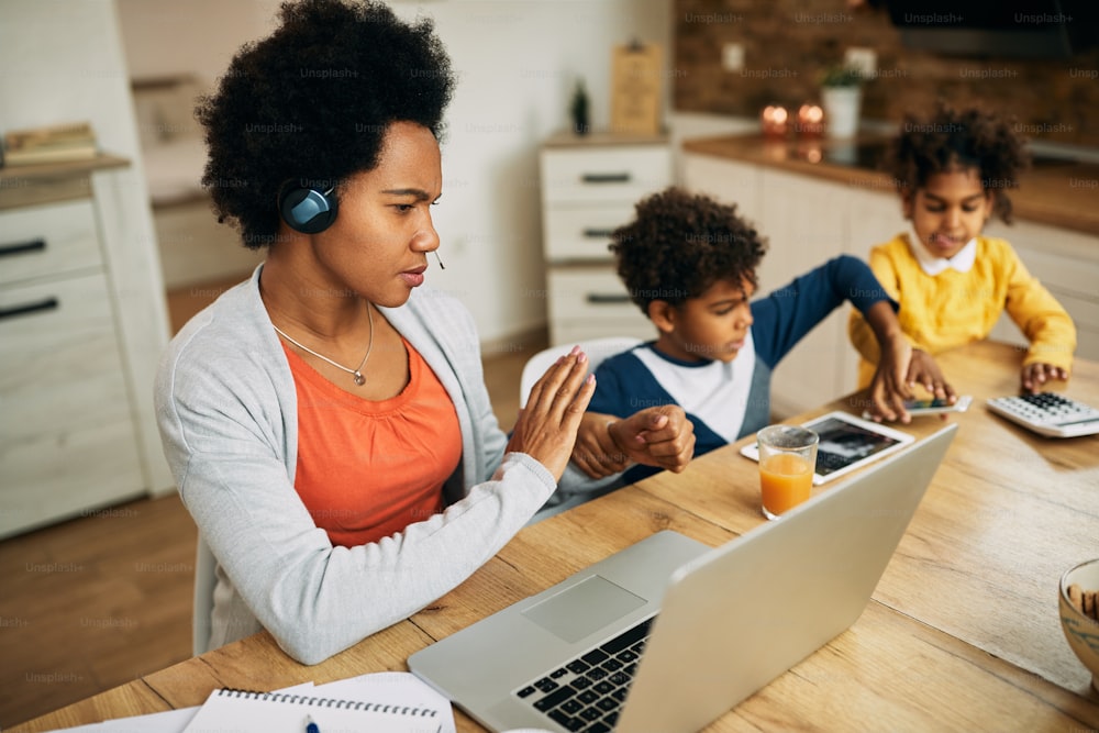 African American working mother silences her children while having conference call over laptop at home.