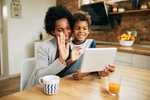 Madre e hijo negros felices usando el panel táctil y saludando a alguien mientras tienen una videollamada en casa.
