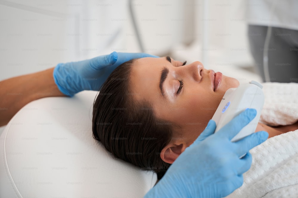Woman face receiving ultrasound procedure in cosmetology office. Hands in gloves of professional doctor dermatologist stroking with cosmetic apparatus skin of woman