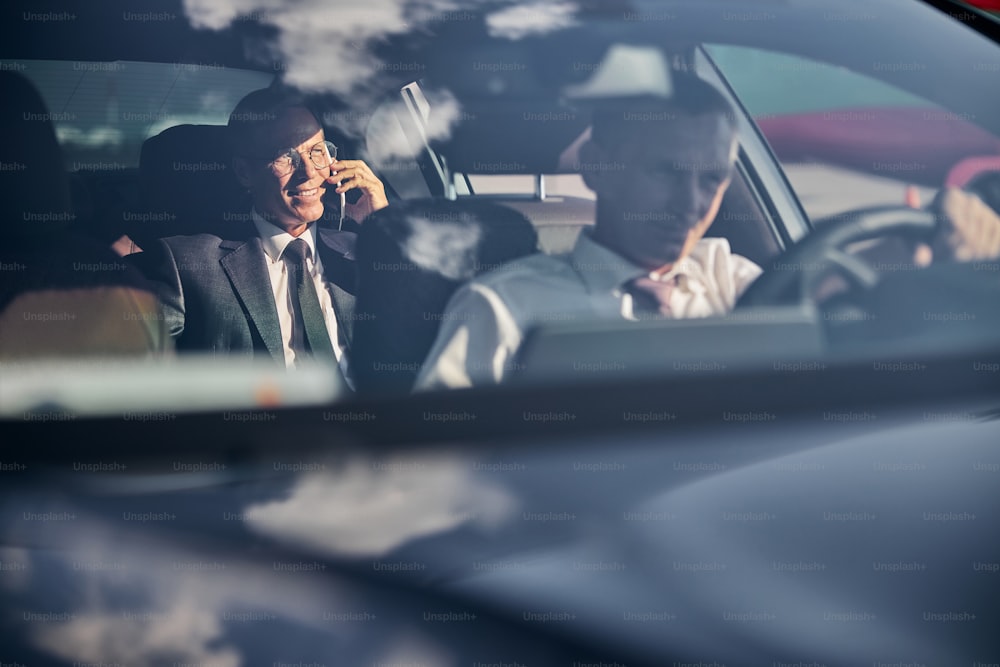 Hombre está conduciendo un automóvil cómodo mientras su jefe está hablando por teléfono celular en la parte de atrás