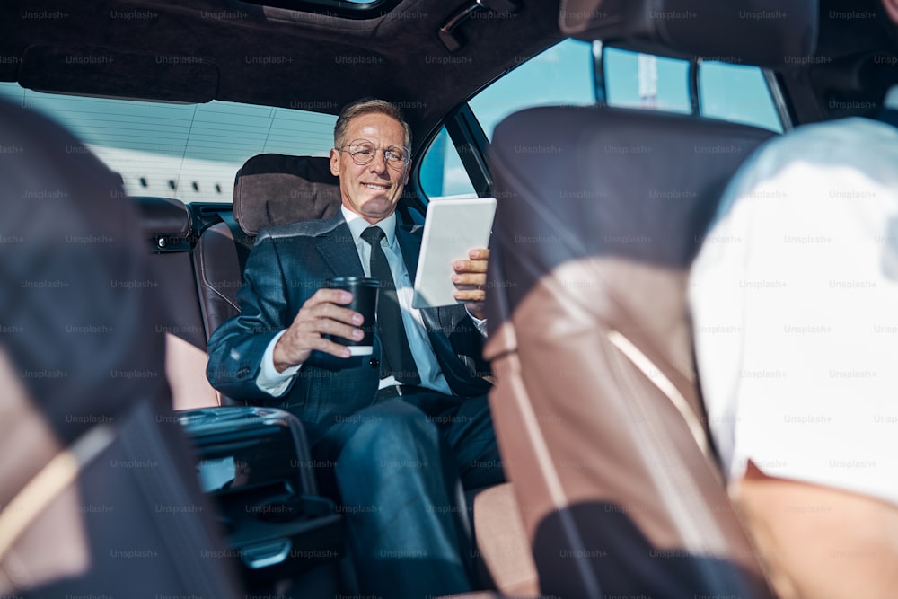 Fröhlicher, eleganter Mann sitzt nach der Ankunft von der Reise hinten im Auto mit Touchpad und Tasse Kaffee