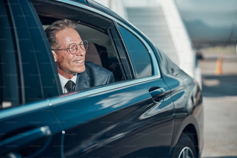 Cheerful elegant man in glasses is looking after automobile window during transfer after landing by plane