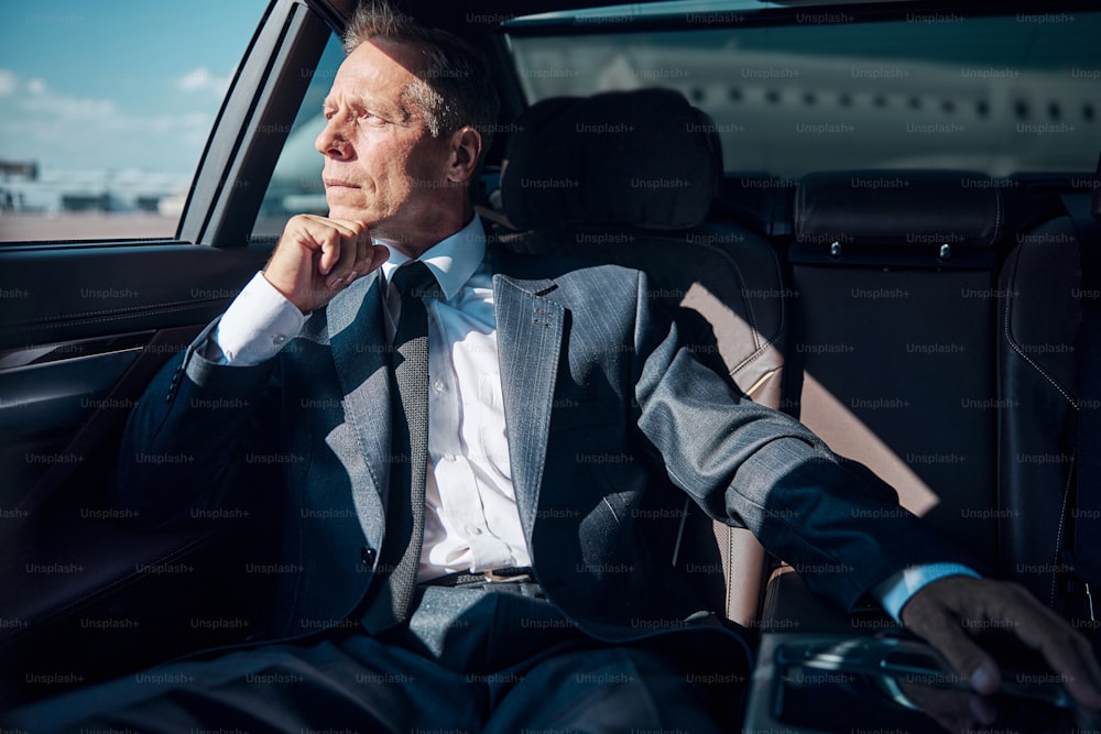 Pensive mature man in elegant suit is sitting on back seat after landing by plane
