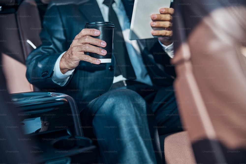 Cabeza recortada de hombre elegante en traje y corbata sentado en la parte posterior con taza de bebida caliente y panel táctil durante la transferencia