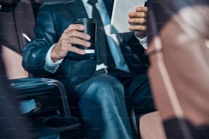 Cropped head of elegant man in suit and tie sitting in back with cup of hot drink and touchpad during transfer