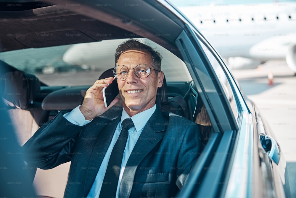 Hombre maduro alegre con ropa elegante se comunica por teléfono celular mientras sale del aeropuerto en automóvil