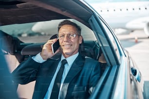 Cheerful mature man in elegant wear is communicating on cell phone while leaving airport by car