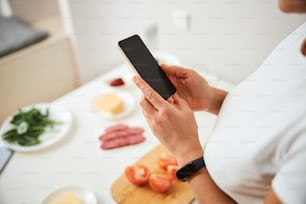 Cropped photo of a smartphone in hands of a woman standing by the kitchen table. Copy space on the screen