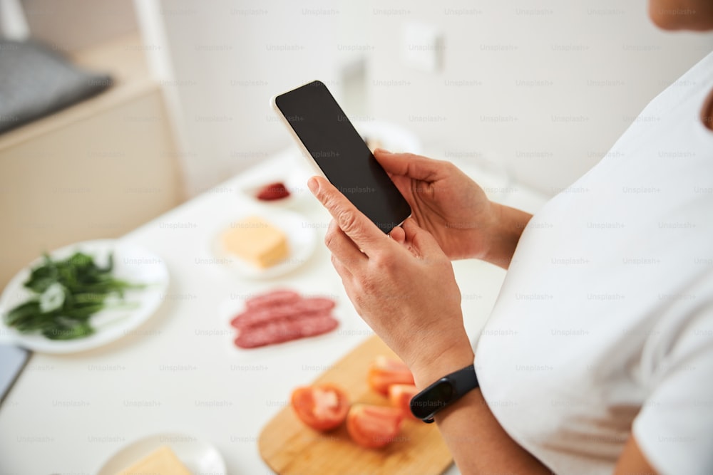 Photo recadrée d’un smartphone dans les mains d’une femme debout près de la table de la cuisine. Copier l’espace à l’écran