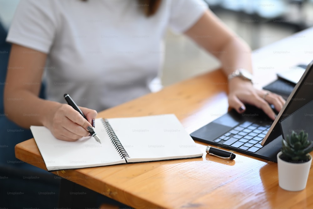 Close up view of female using computer tablet and making notes or planning schedule in notebook.