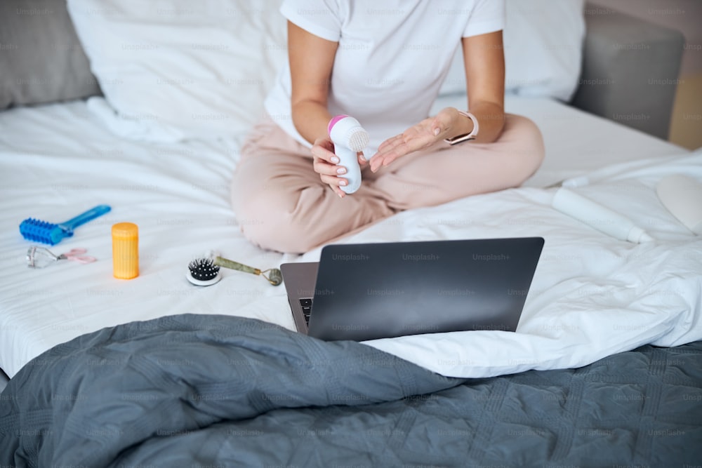 Cropped head portrait of unrecognized female in white shirt holding cleaning massager in hand while sitting in front of laptop
