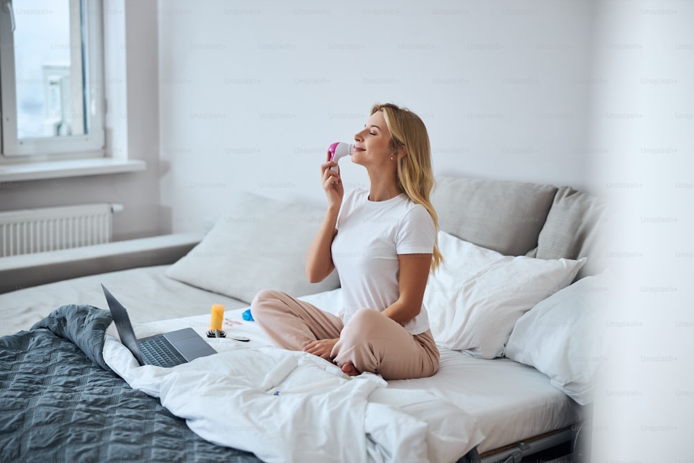 Full length side view portrait of pretty elegant lady in home clothes sitting on bed near laptop while using cleaner massager in home apartments