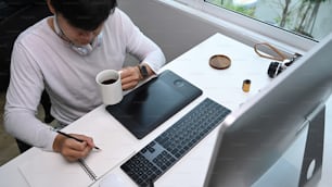 Über Kopfaufnahme eines Grafikdesigners, der Ideen in ein Notizbuch schreibt und am Arbeitsplatz Kaffee trinkt.
