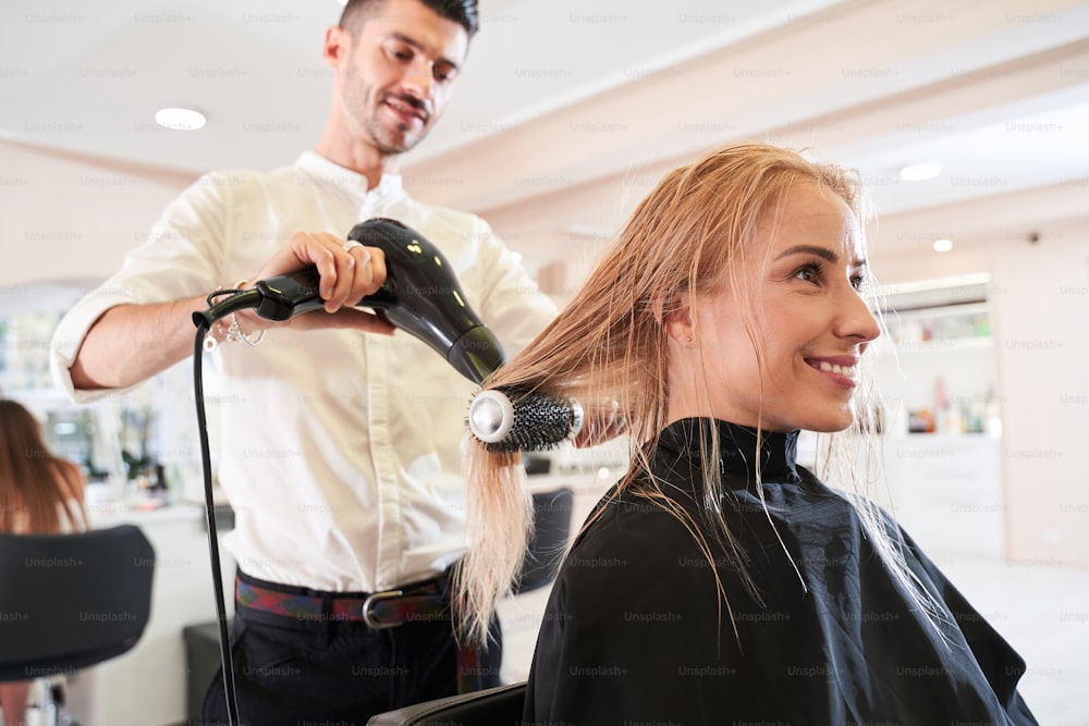 Young unshaven male hair stylist using blower and brush to dry hair. Woman at a sophisticated beauty parlor getting a hairdo. Stock photo