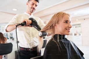 Young unshaven male hair stylist using blower and brush to dry hair. Woman at a sophisticated beauty parlor getting a hairdo. Stock photo