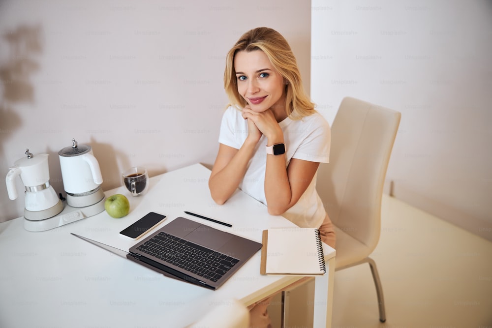 Waist up portrait of cute blonde female sitting at the table while working on the laptop in home office