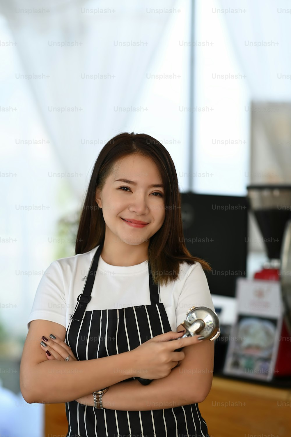 Portrait of cheerful women baristas wearing aprons standing at the counter in cafe and holding coffee making accessory.