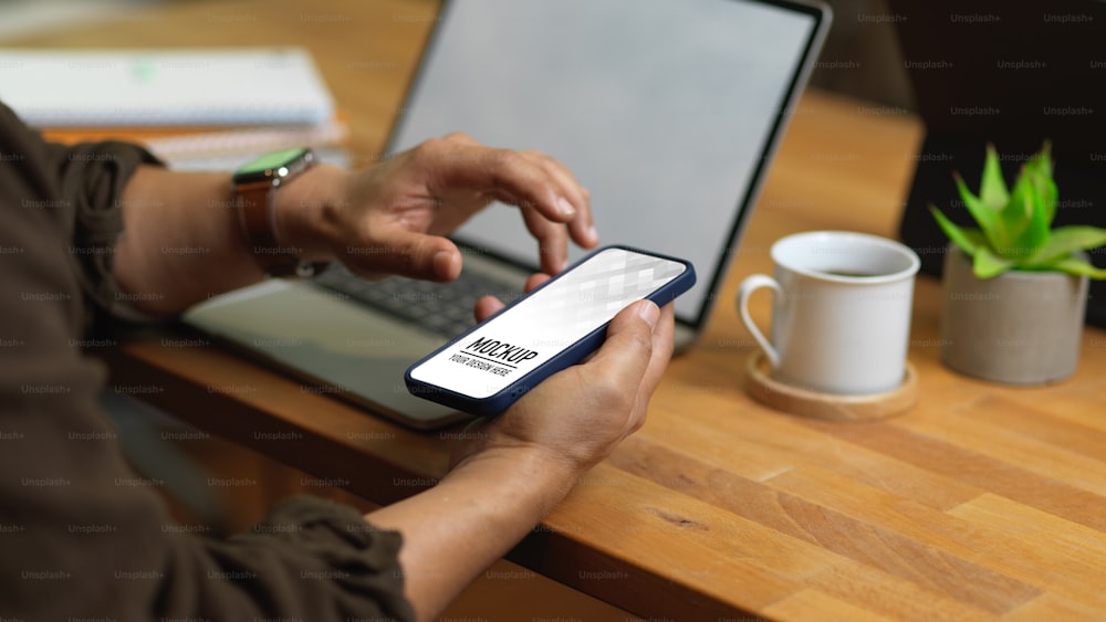 Cropped shot of male hands using smartphone on worktable with laptop in office room