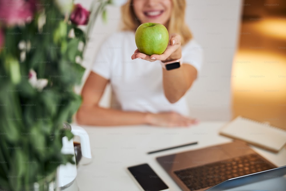 Close up portrait of beautiful elegant woman with fresh fruit in arm sitting at the laptop while posing at the photo camera in room indoors