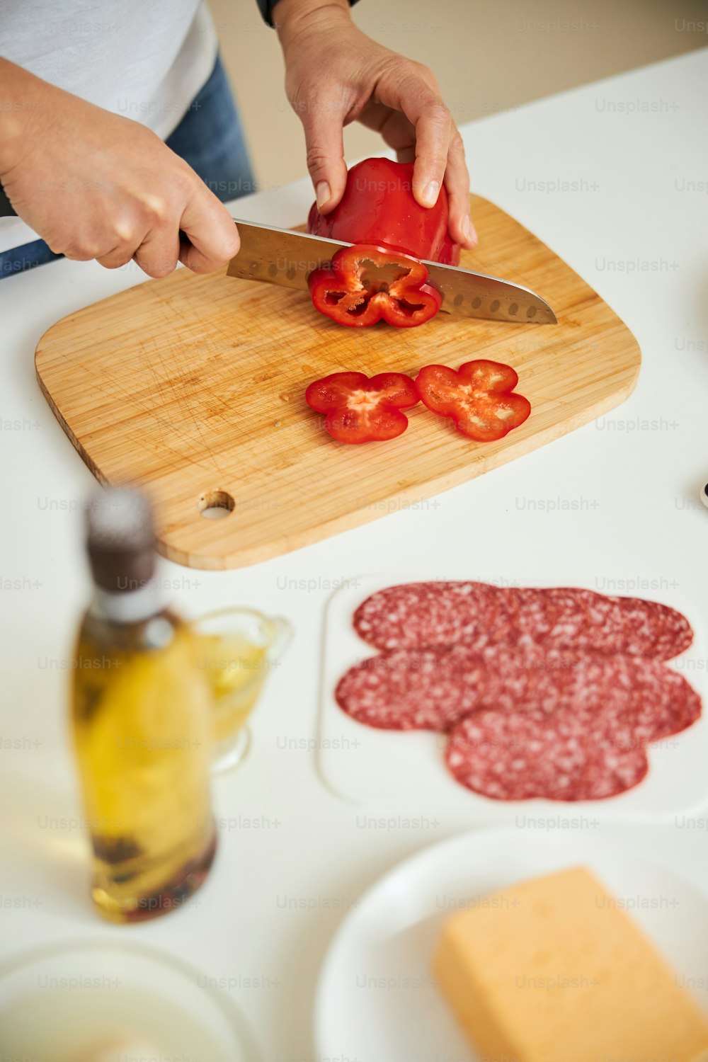 Close up of a kitchen table with slices sausages and a bottle of oil. Woman cutting pepper on the board