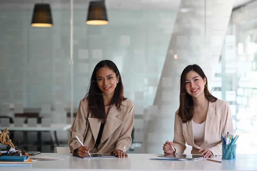 Deux collègues femmes d’affaires joyeuses travaillant ensemble sur un nouveau projet et souriant à la caméra dans un bureau moderne.