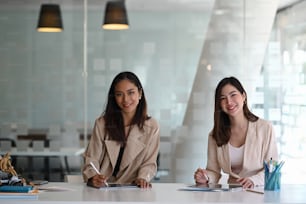 Two cheerful businesswoman colleagues working together on new project and smiling to camera in modern office.