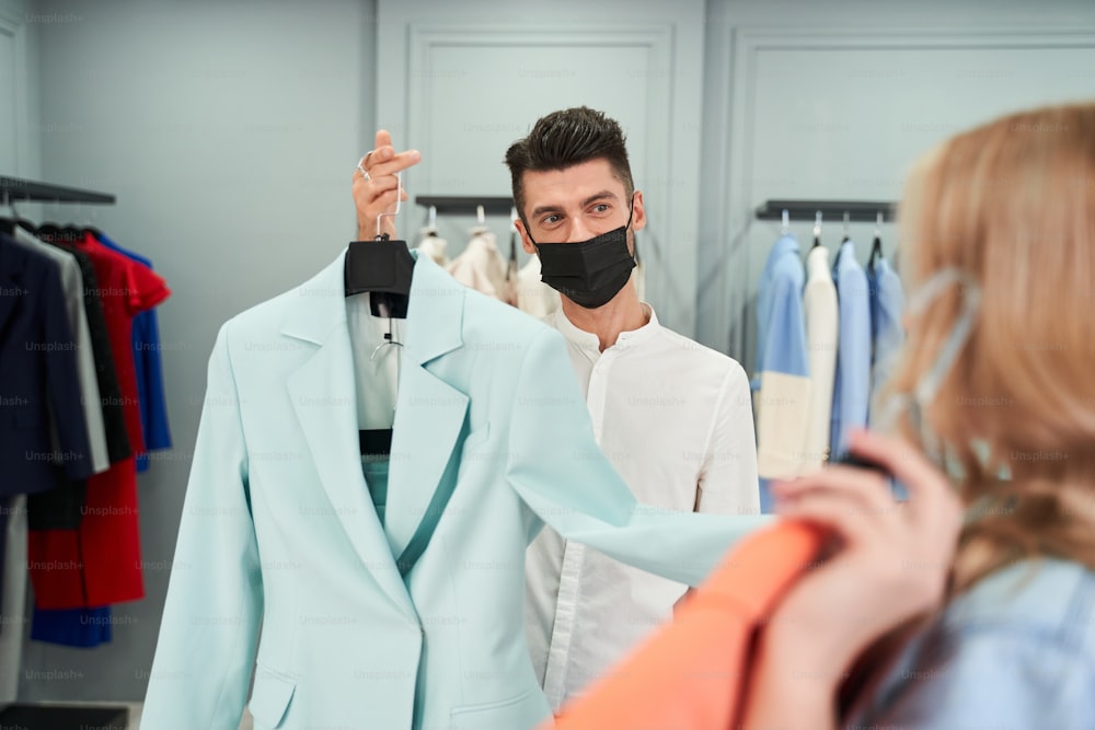 Having a new purchase. Thoughtful young woman choosing clothes from the rack while standing in the showroom with assistant wearing protective mask