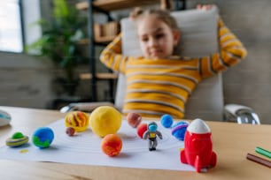 8 years old girl study Solar system sitting on the desk in the room. Female elementary school student doing her homework - sculpts planets models from kids clay or plasticine. Selective focus on the planets.