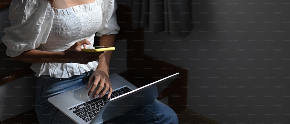 Mulher freelancer nova que trabalha no laptop e usa o telefone inteligente enquanto está sentada na escada em casa.