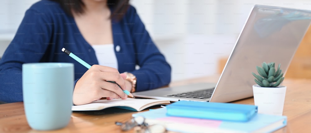 Estudiante universitaria joven que usa una computadora portátil que estudia en línea desde casa y toma notas en el cuaderno.
