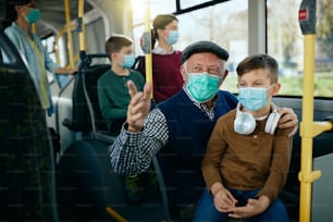 Glücklicher älterer Mann, der mit seinem Enkel spricht, während er mit dem Bus pendelt und Gesichtsmasken während der COVID-19-Pandemie trägt.
