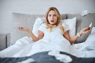 Attentive blonde being at a loss while checking her temperature with electro thermometer