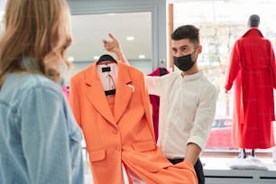 What do you think about this one? Young caucasian stylist or assistant wearing protective mask helping to choose clothes to his customer while showing an orange suit to the woman