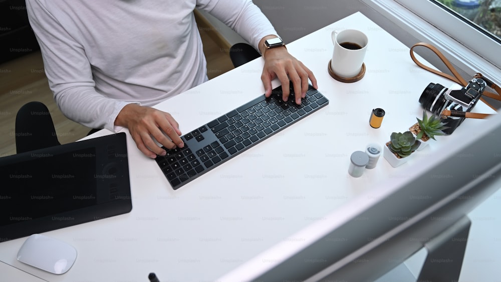 Foto aérea de mãos de fotógrafo jovem digitando no teclado em seu espaço de trabalho.