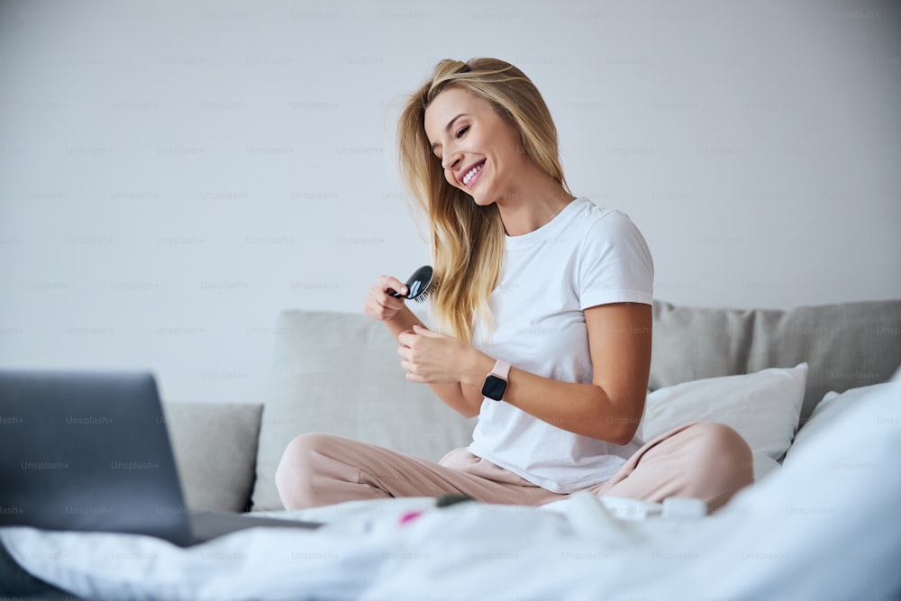 금발 백인 매력적인 여성의 측면보기 초상화는 노트북에서 무언가를 보고 홈 아파트에서 머리를 빗는 동안 침대에서 시간을 보내고 있다