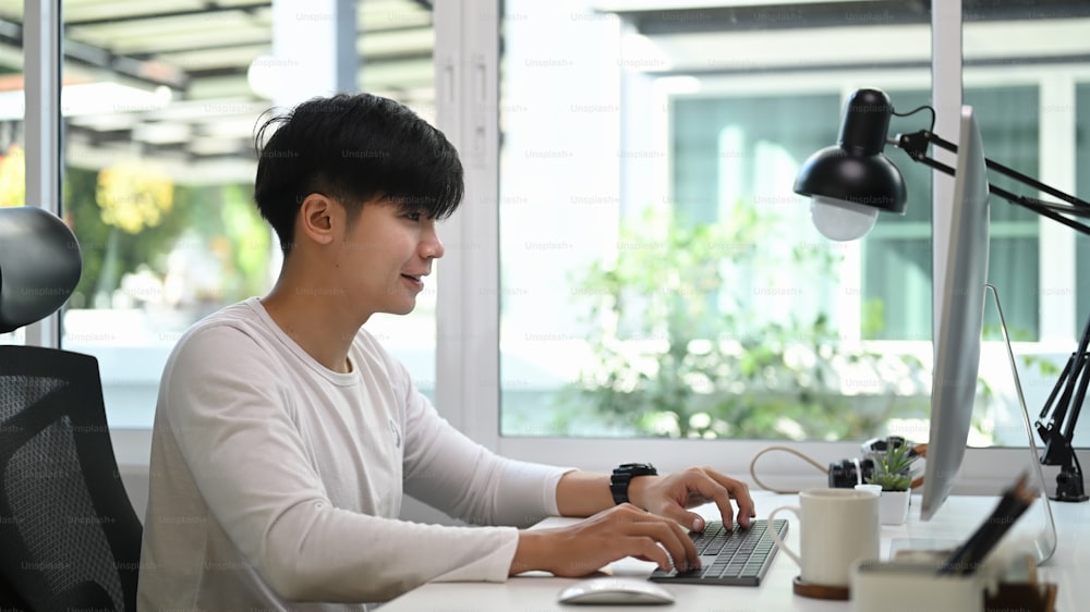 Vista lateral do homem freelancer em casualwear sentado em home office e trabalhando em projeto com computador.