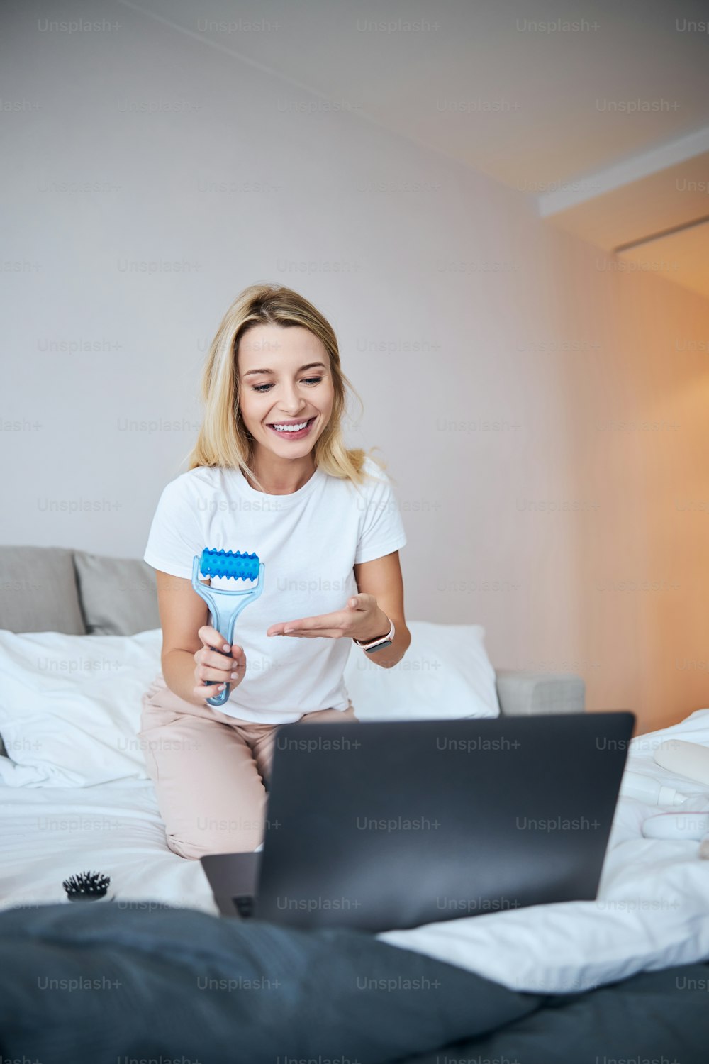 Close up portrait of happy smiling female in white shirt talking on video call with friends and demonstrating blue roller massager in room indoors