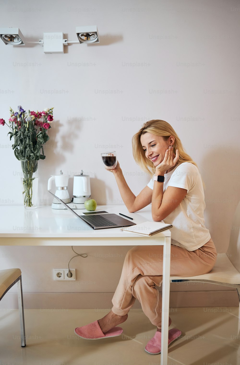 Happy smiling blonde woman in white shirt with smart watch on hand holding cup of coffee in hand and smiling in the kitchen