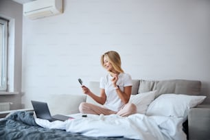 Full length side view portrait of happy cheerful female in home clothes holding brush in hand while resting in the bed in the morning