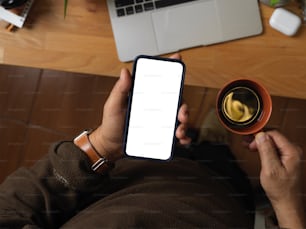 Vista superior da mão masculina segurando o smartphone e a xícara de café acima da mesa de trabalho