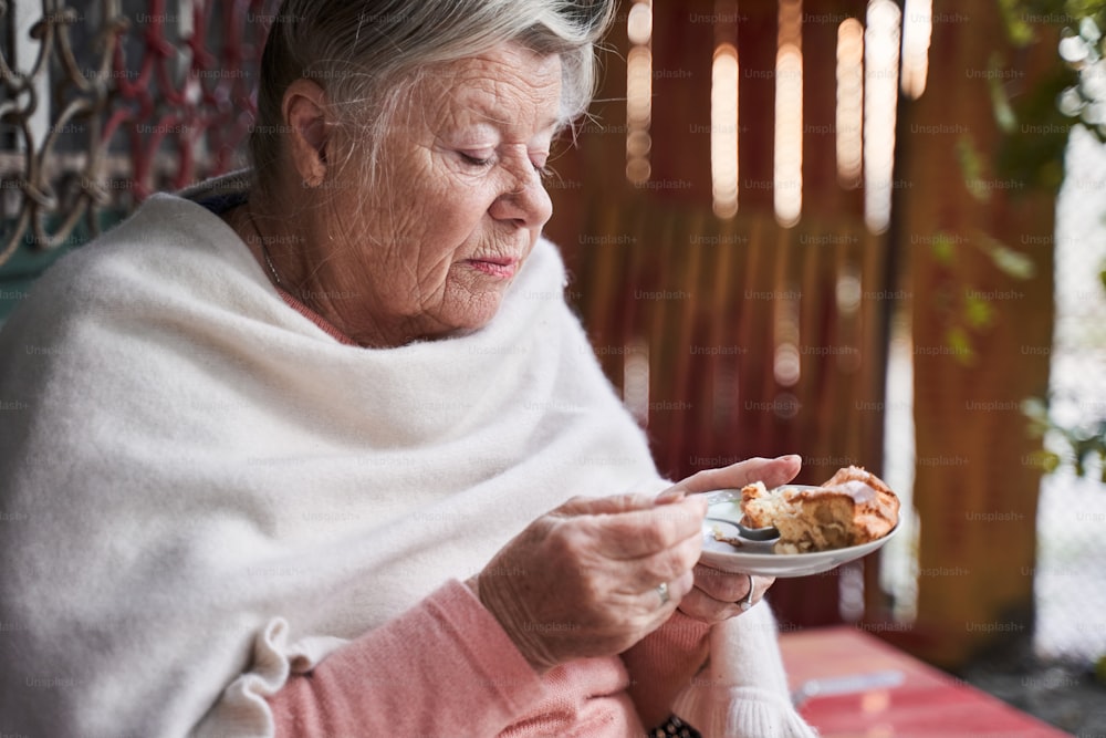 Kind elderly woman eating pie from the plate with appetite while sitting on the bench near the country house. Stock photo