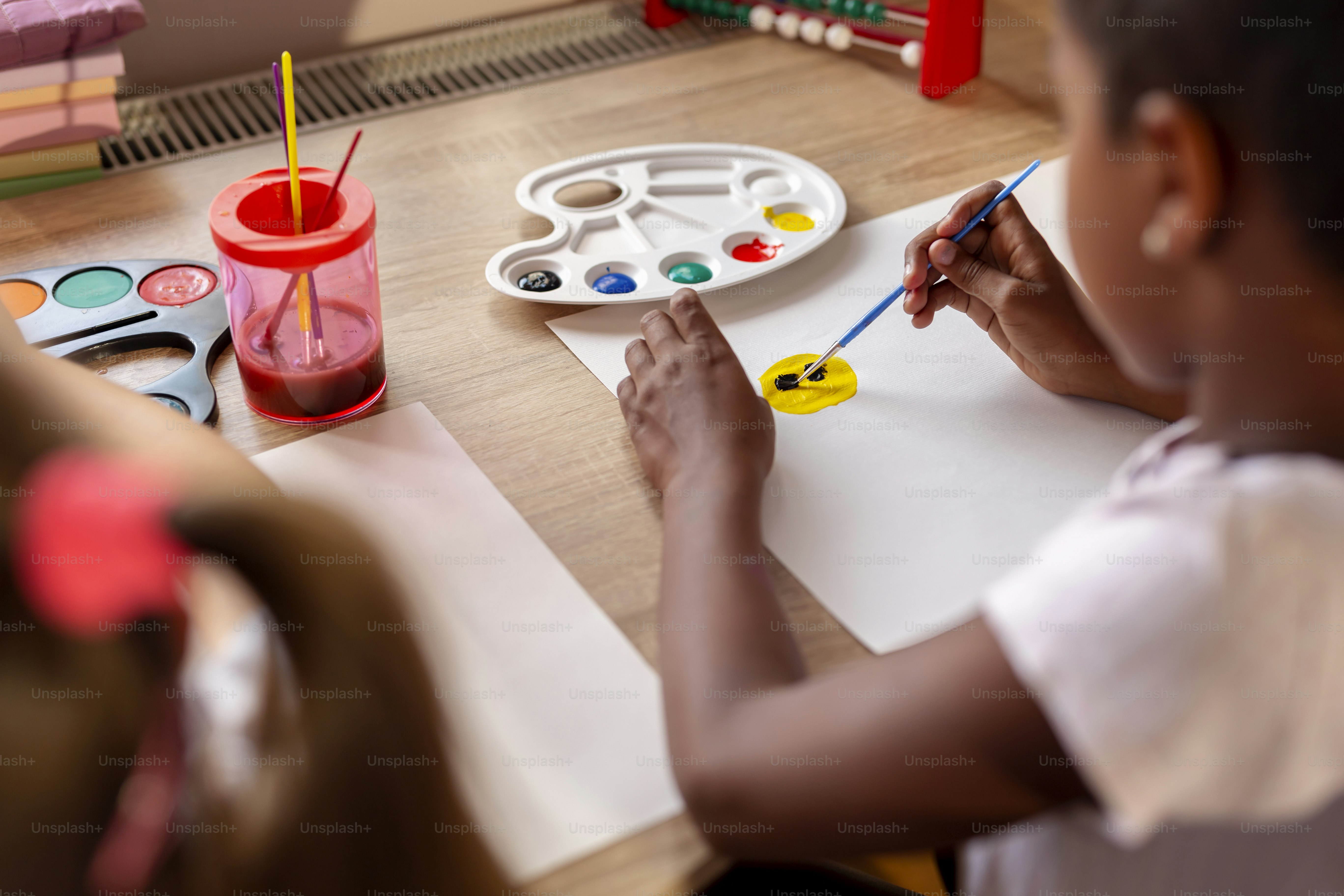 Two little girls sitting at a desk, painting with water colors and having fun while doing homeschooling art class due to Covid 19 pandemic social distancing measures