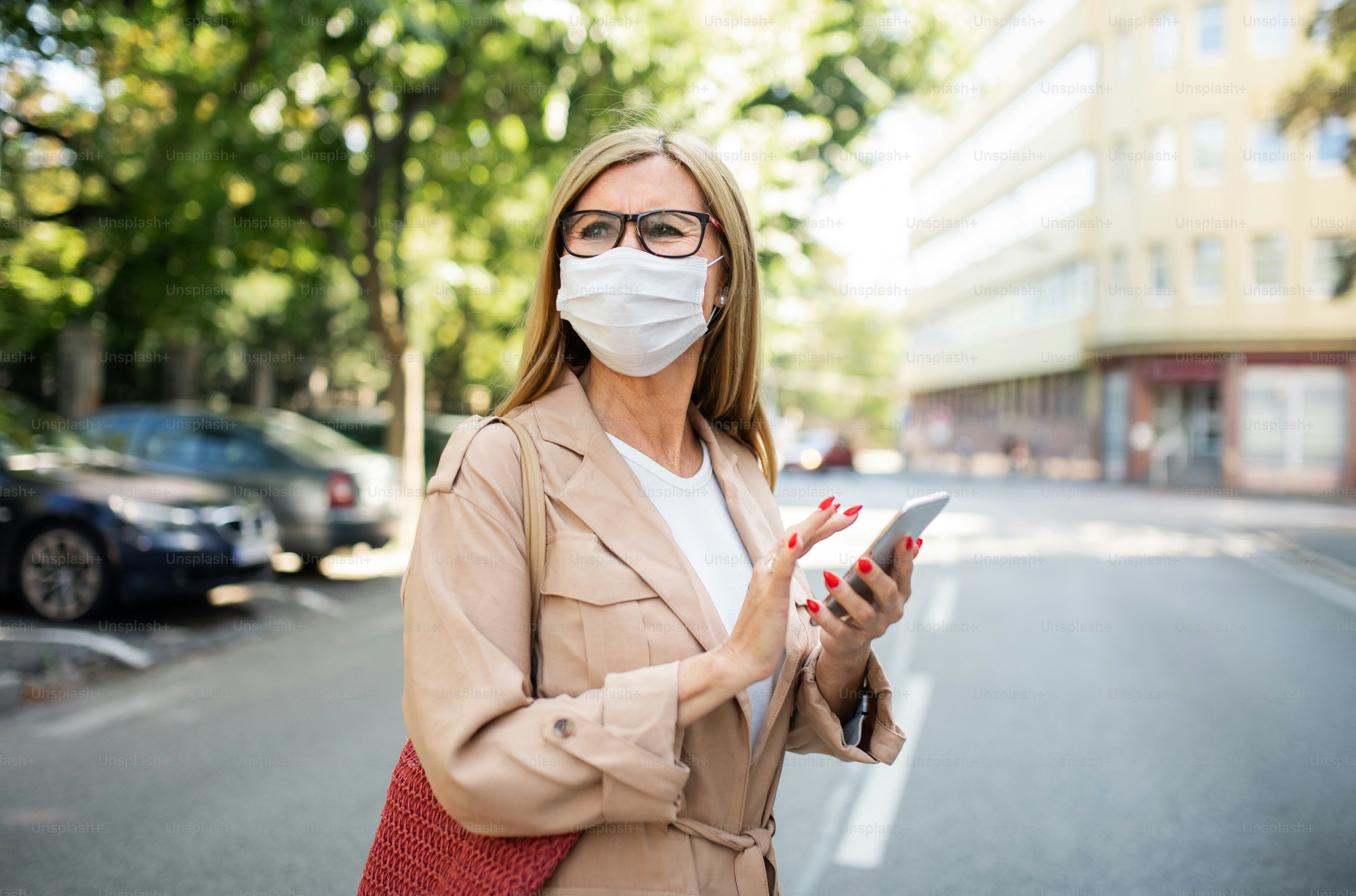 Portrait of senior woman with smartphone crossing street outdoors in city or town, coronavirus concept.