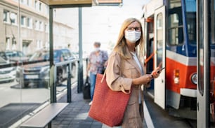Portrait of senior woman with smartphone on bus stop outdoors in city or town, coronavirus concept.