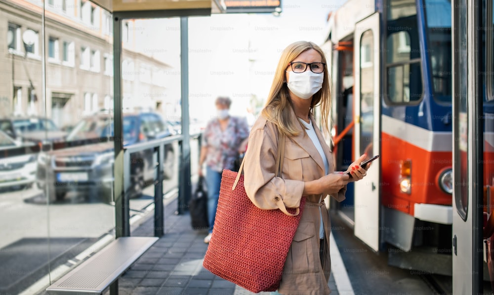 Retrato de una mujer mayor con un teléfono inteligente en una parada de autobús al aire libre en la ciudad o pueblo, concepto de coronavirus.
