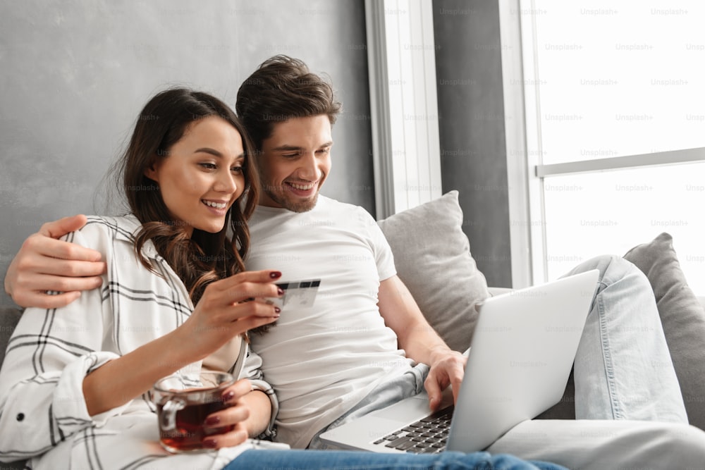 Encantadora pareja de hombre y mujer sentados en el sofá en el apartamento cerca de la ventana grande mientras usan la computadora portátil y la tarjeta de crédito para comprar en línea